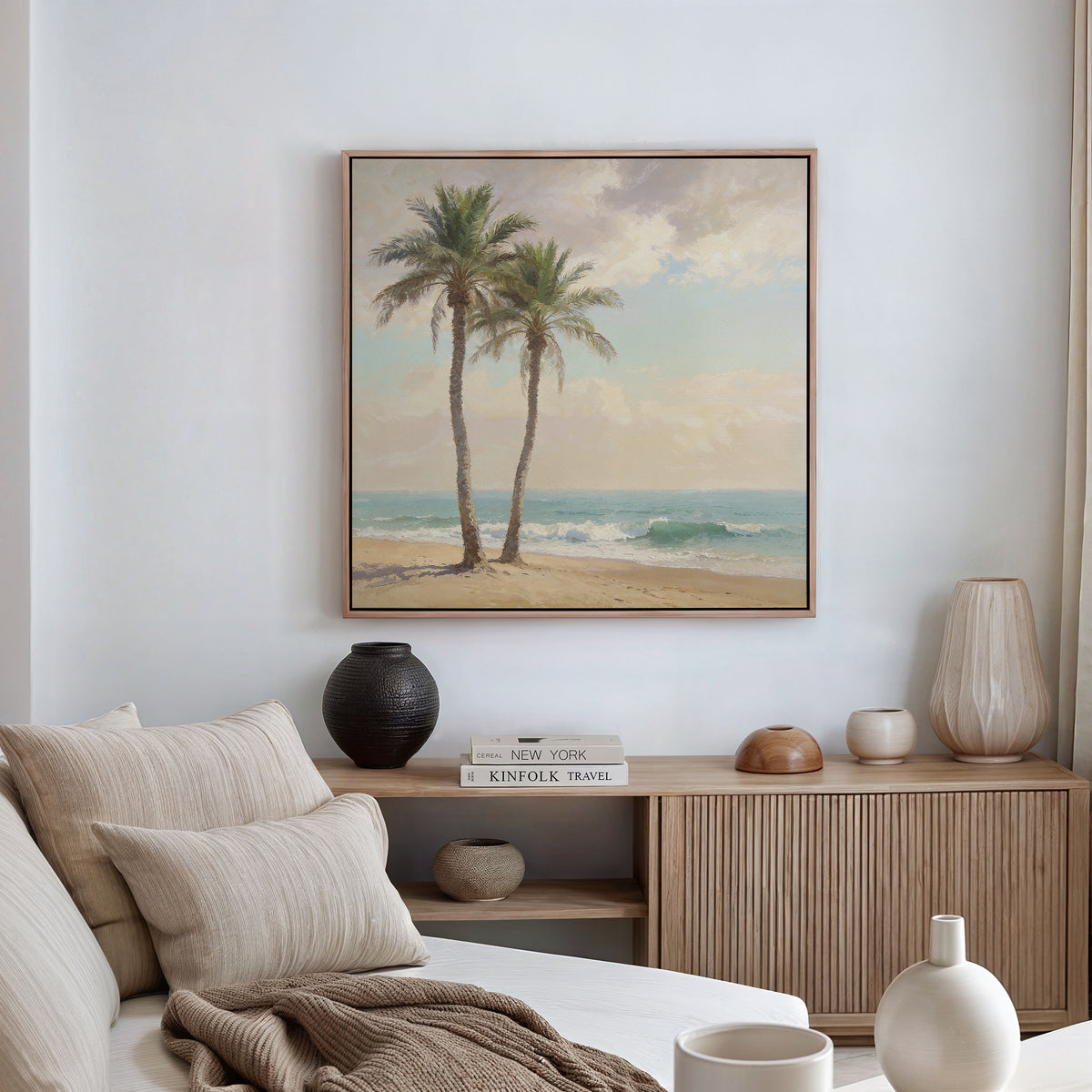A serene living room with a beige sofa, striped cushions, and a wooden console topped with vases and books. The Paradise Found Canvas Art - Square featuring palm trees on a beach hangs on the white wall above.