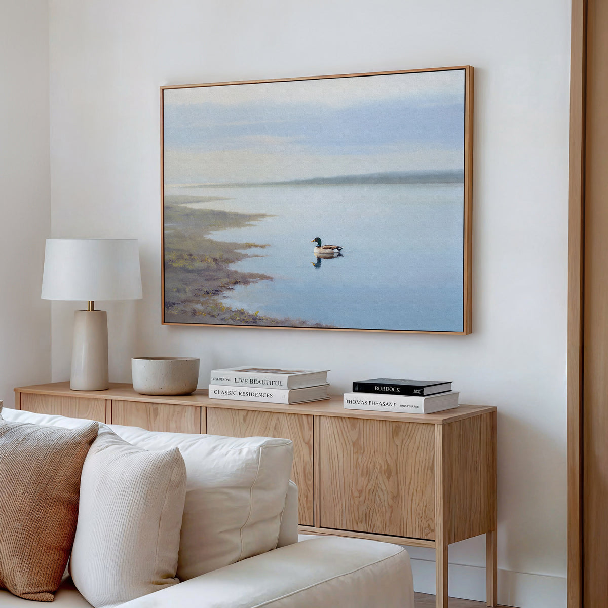 A minimal living room with a light wooden sideboard, books, a lamp, and the Peaceful Duck Haven Canvas Art - Horizontal above a white sofa creates a tranquil Nordic Park vibe.