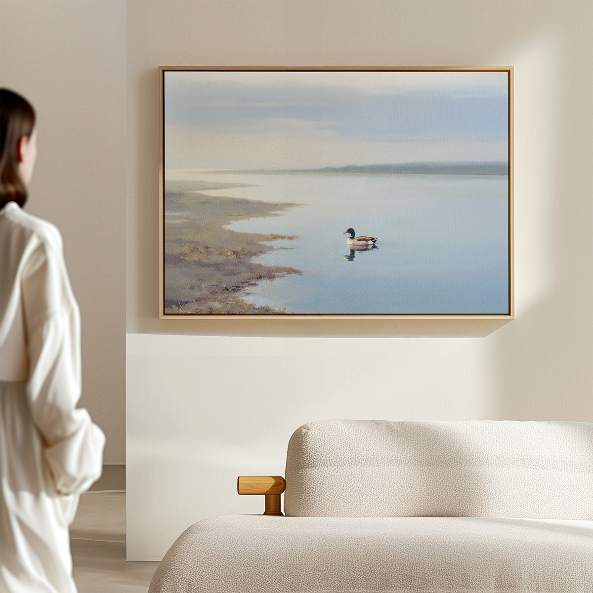 A woman stands in a softly lit room, admiring the Peaceful Duck Haven Canvas Art - Horizontal, featuring a tranquil duck on calm water, displayed above a beige sofa.