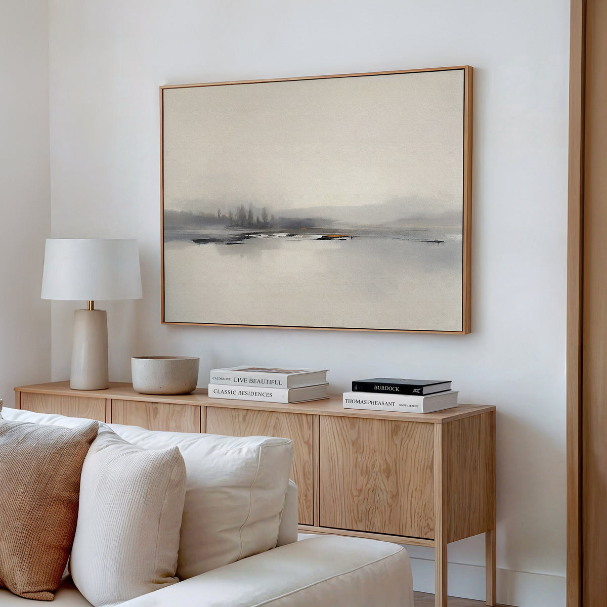 A modern minimalist living room features a light wood sideboard, white lamp, books, and bowl beneath the Peaceful Lake Morning Canvas Art - Horizontal, hanging above a beige couch with cushions to create a peaceful atmosphere.