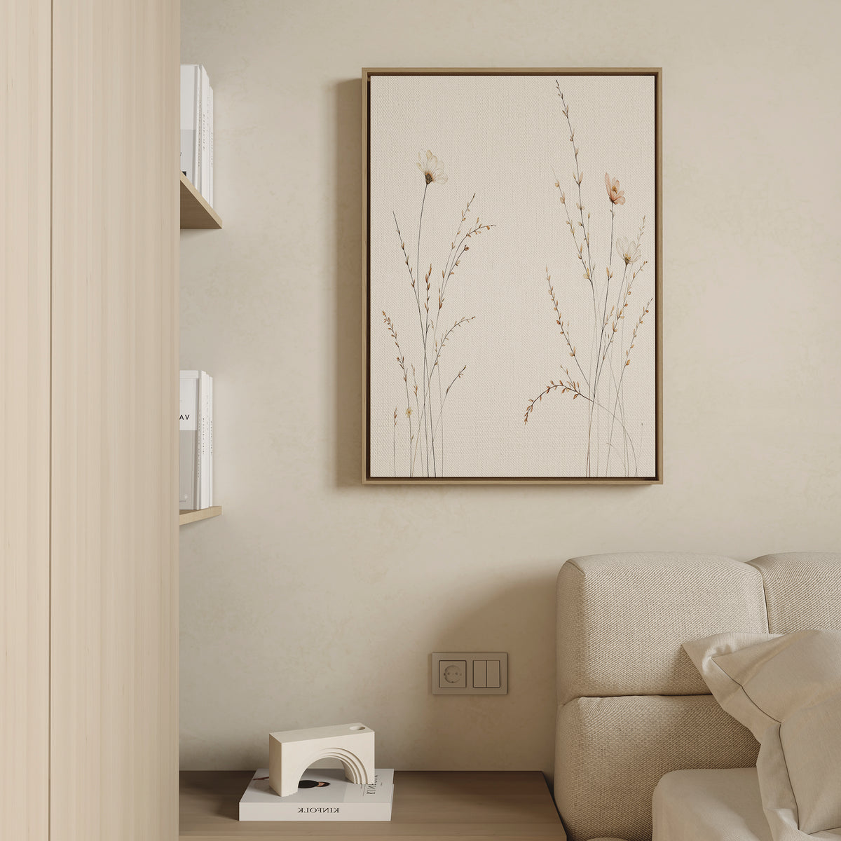 A minimalist living room corner features a beige sofa, the “Prairie Whispers Canvas Art - Vertical” with delicate grasses on the wall, shelves of white books, and a small geometric decor piece for an atmosphere of natural tranquility.