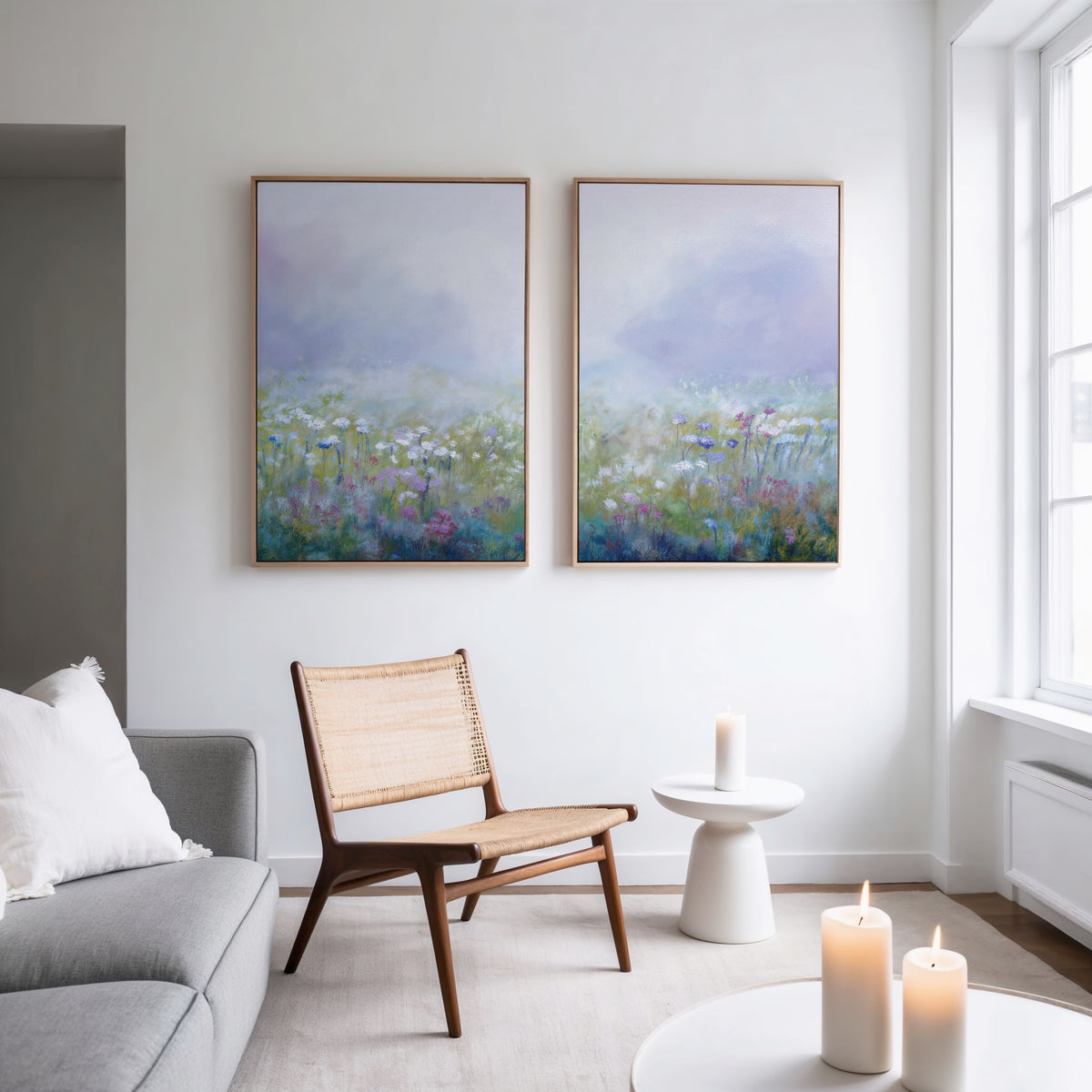 A modern, minimal living room has a gray sofa, woven chair, small white table, three lit candles, and the "Purple Haze - Set of 2" framed canvas prints on a white wall near a large window.