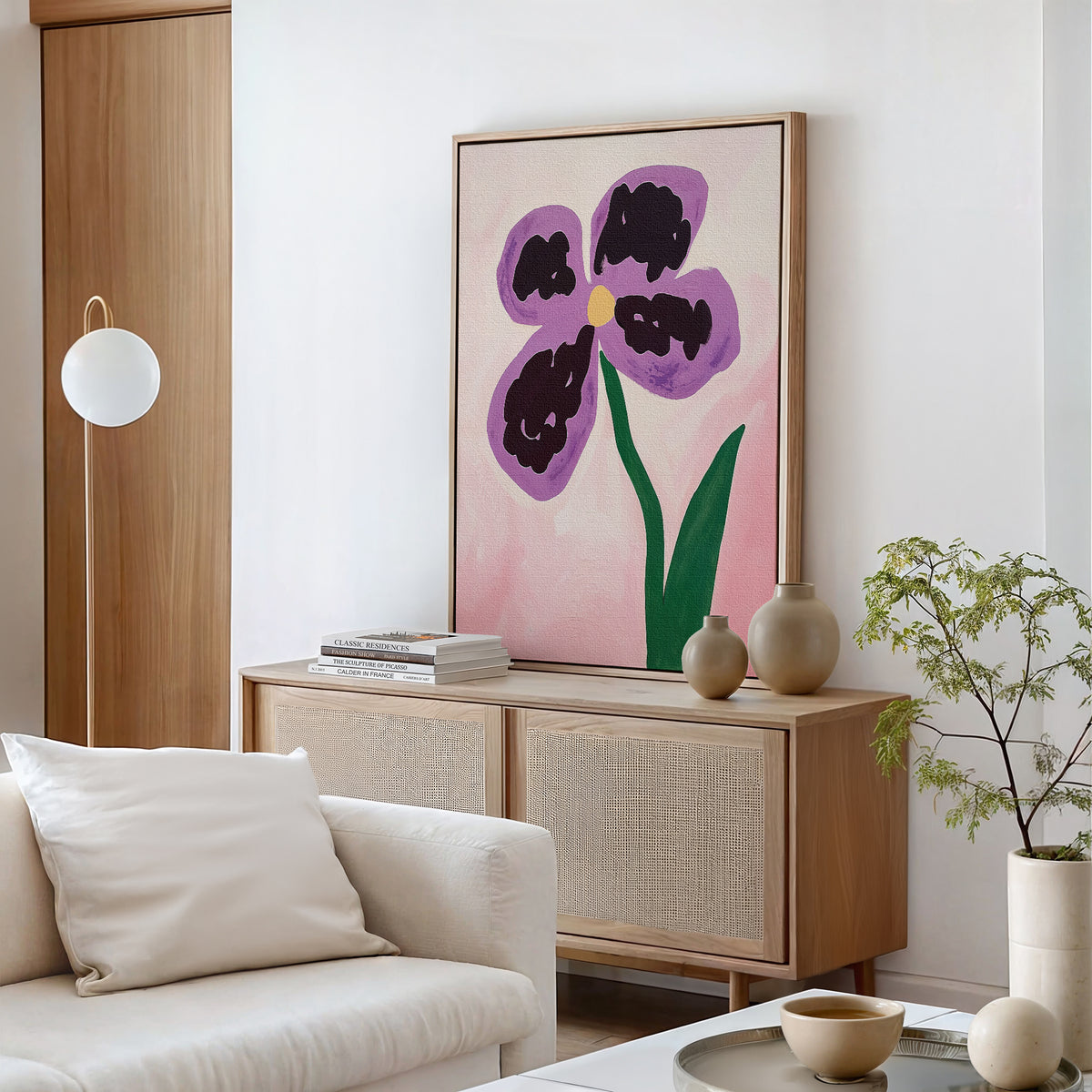 A modern living room with a white sofa, light wood sideboard, and the Purple Pansy Canvas Art - Vertical on the wall. Neutral decor, books, ceramic vases, and a small green plant complete this Nordic Park-inspired space.