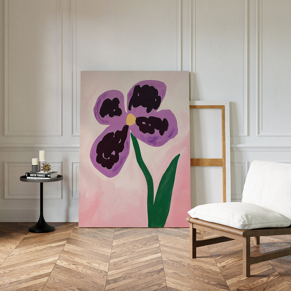The Purple Pansy Canvas Art - Vertical rests against a white paneled wall in a minimalist room, accompanied by a white cushioned chair, an empty frame, and a small round table holding candles and books.