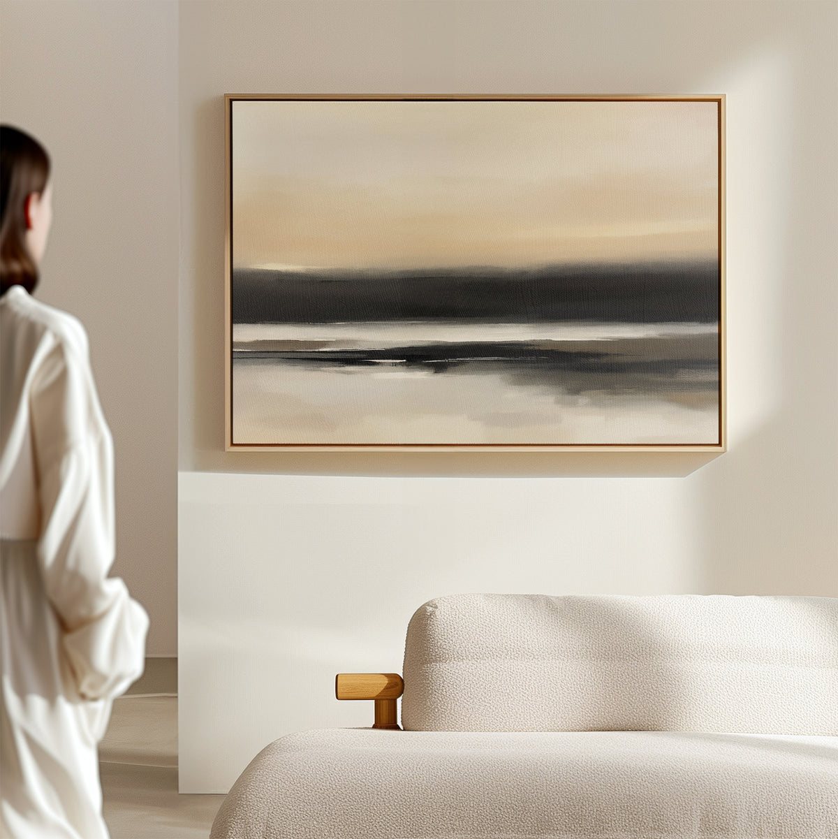 A person in a white outfit stands in a minimal room, gazing at the Quiet Shoreline Canvas Art - Horizontal, an abstract print with soft beige, gray, and black tones hanging above a light-colored sofa.