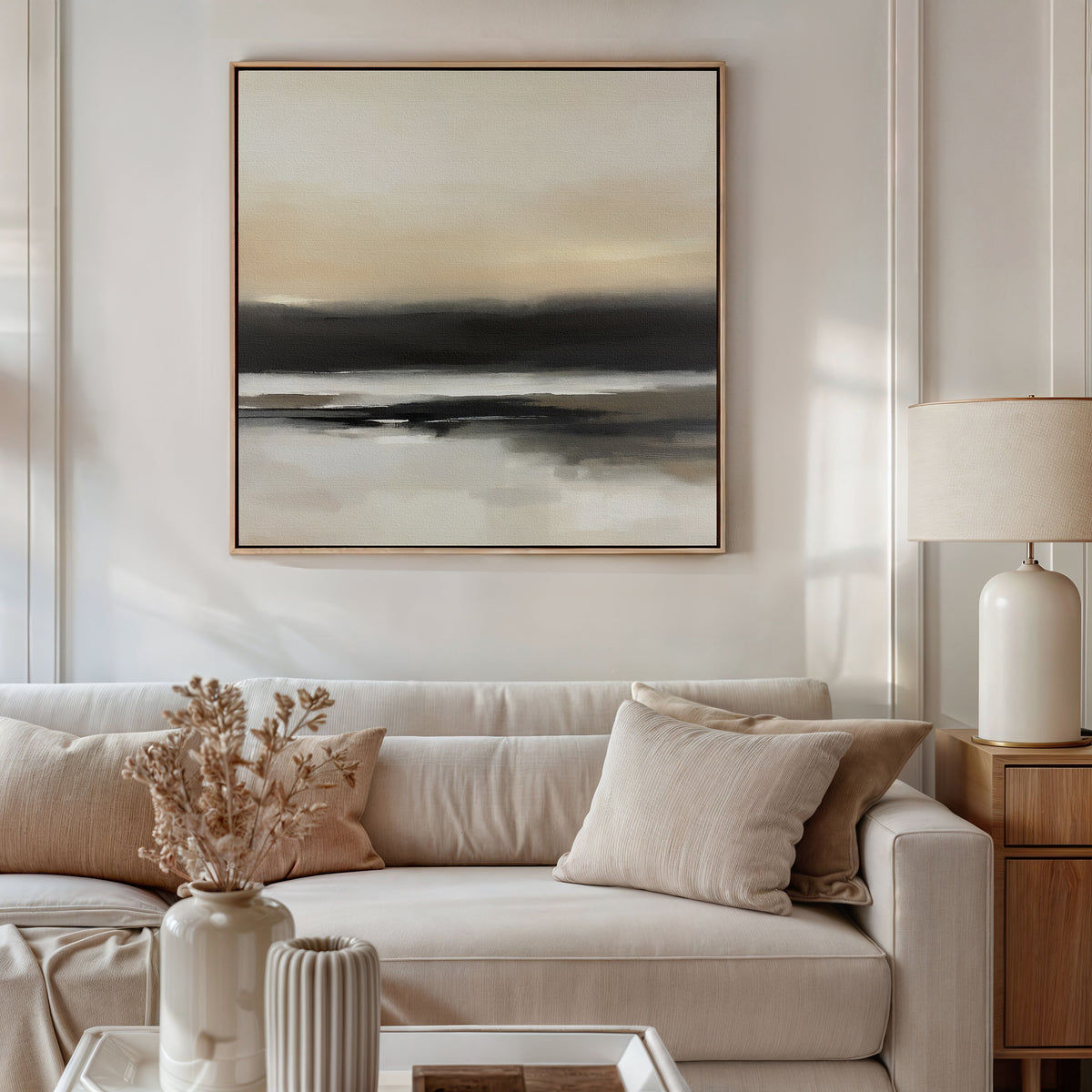 A modern living room features a beige sofa, light cushions, minimalist decor, a wooden side table with a lamp, and the Quiet Shoreline Canvas Art - Square in neutral tones. A vase with dried flowers sits on the coffee table.