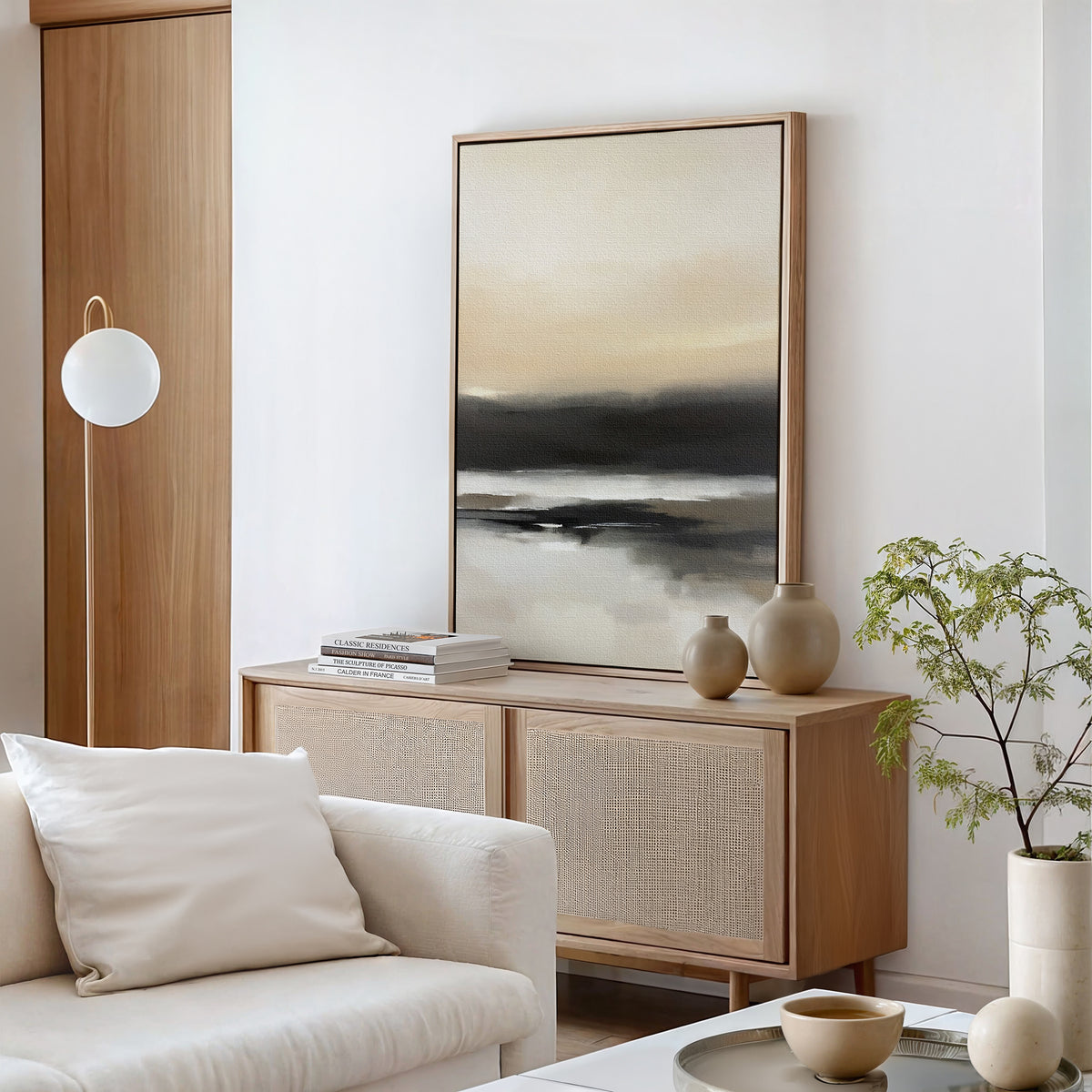 A modern living room features the Quiet Shoreline Canvas Art - Vertical above a light wood sideboard, paired with neutral vases, a white sofa with cushions, a round floor lamp, and a small green plant.