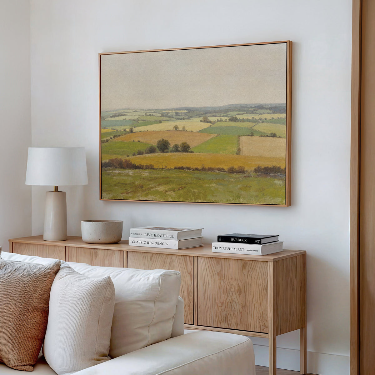 A cozy living room with a light wood sideboard, neutral decor, and the Rolling Fields Canvas Art - Horizontal above. A lamp, bowl, and stacked books rest on the sideboard beside a white sofa.