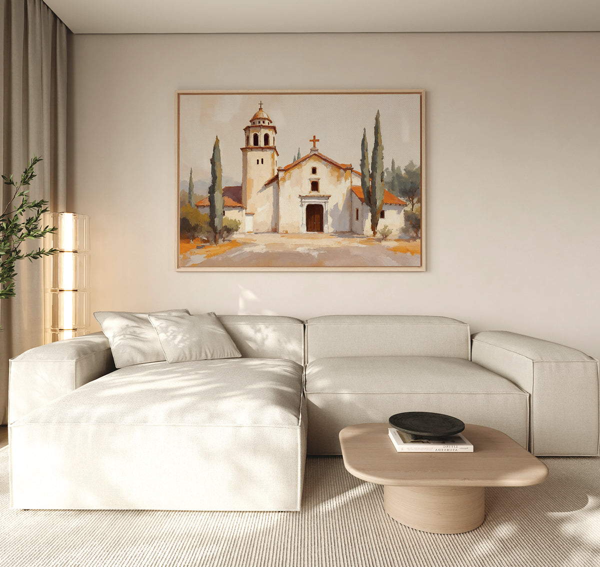 A modern living room features a light beige sectional, a round wooden coffee table, and the Sacred Courtyard Canvas Art - Horizontal depicting a Spanish colonial church with trees hanging above the sofa. Soft natural light fills the space.