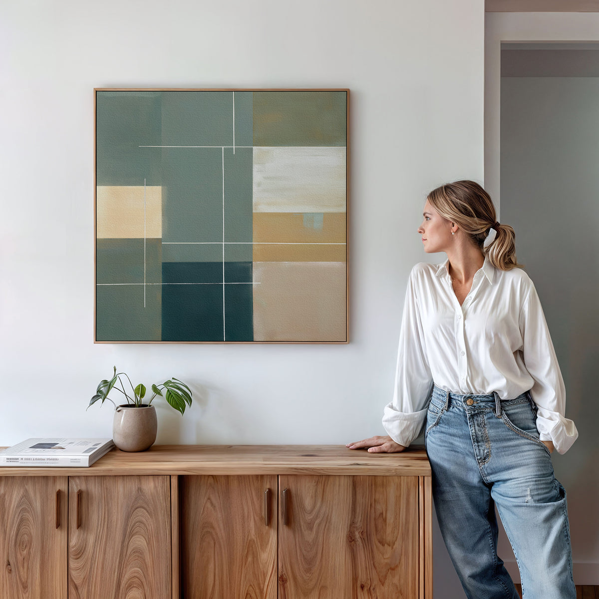 A woman in a white blouse and jeans leans against a wooden cabinet, looking at the Sage Compartments Canvas Art - Square. A small plant and books sit on the cabinet in this minimal, stylish room.