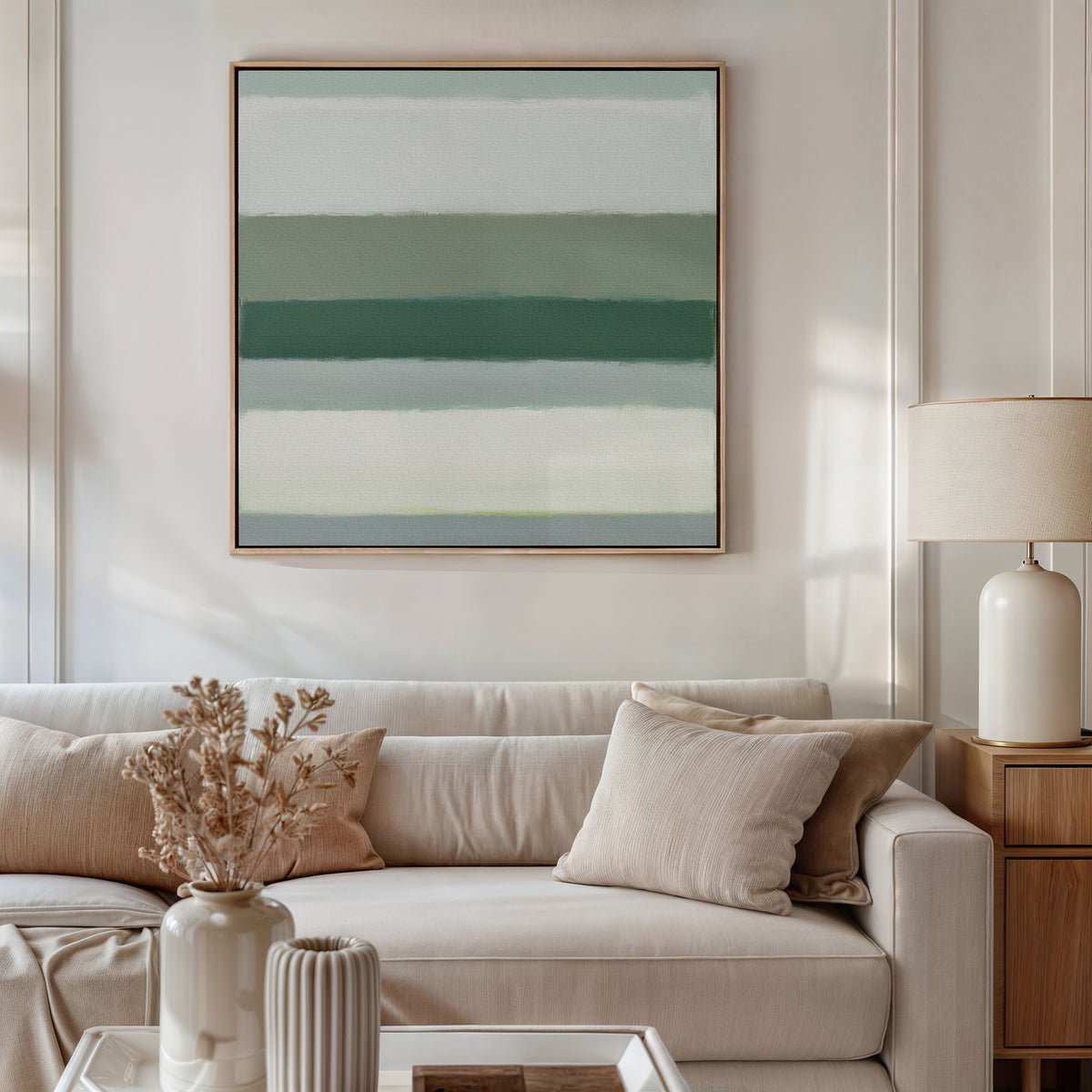 A modern living room features a beige sofa, neutral pillows, and a wooden side table with a white lamp and dried flowers. The wall is adorned with the Sage Gradient Canvas Art - Square, displaying calming green and blue stripes for serene ambiance.
