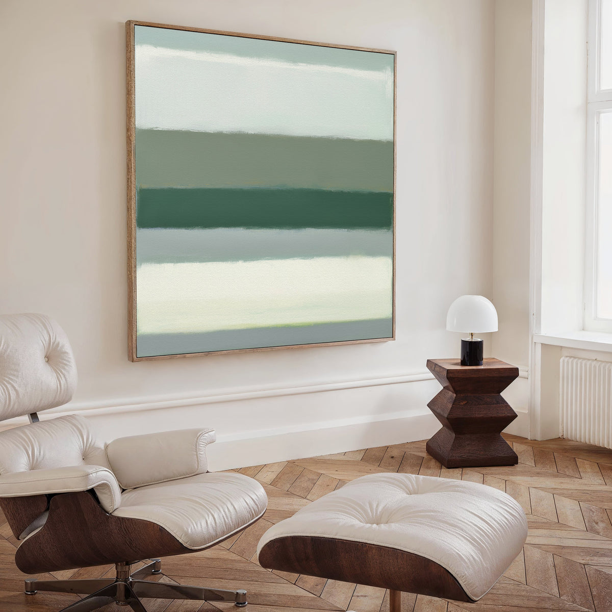 A modern, minimalist living room includes a white leather lounge chair and ottoman, a geometric wooden side table with a white lamp, and the Sage Gradient Canvas Art - Square with soothing green and neutral stripes on the wall.