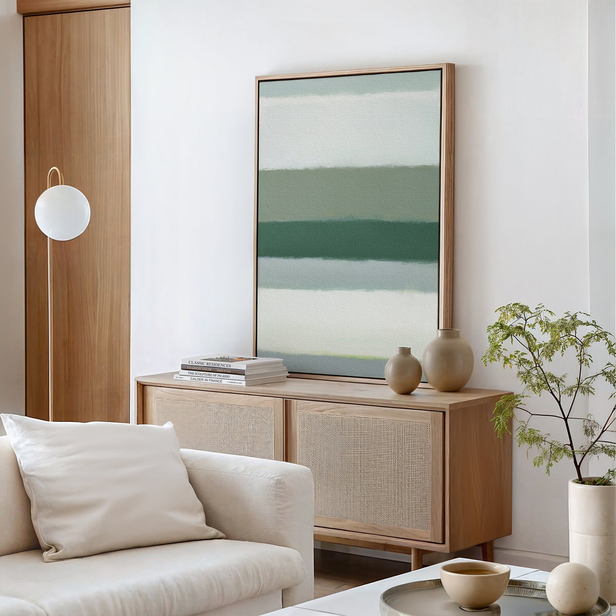 A minimal living room features a light wood cabinet, ceramic vases, books, a potted plant, white sofa with cushions, round hanging lamp, and the Sage Gradient Canvas Art - Vertical for a serene vibe in neutral and green tones.