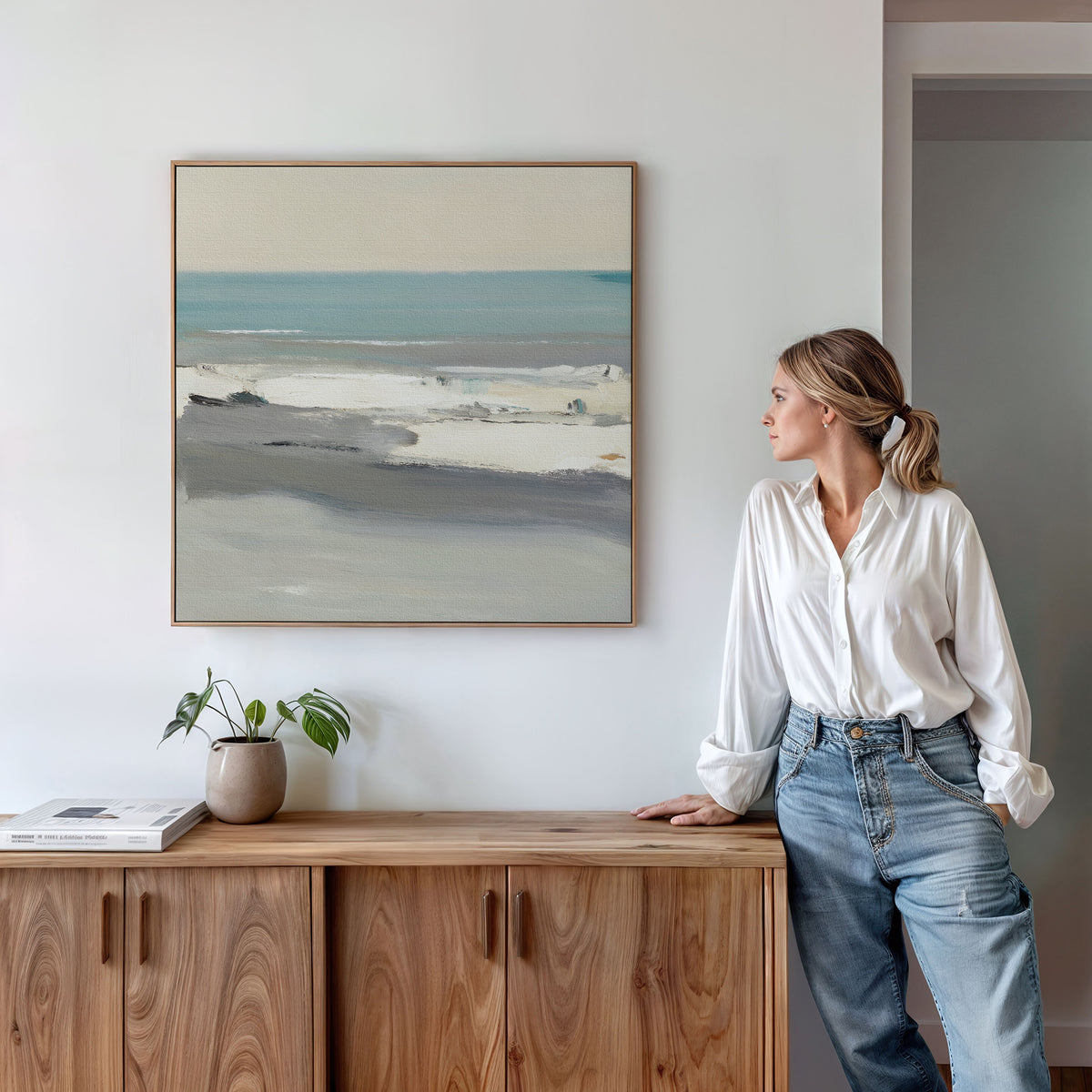 A woman in a white blouse and jeans leans against a wooden sideboard, admiring the Sandy Retreat Canvas Art - Square on a white wall. A plant and book on the sideboard add to the serene seaside atmosphere of the room.