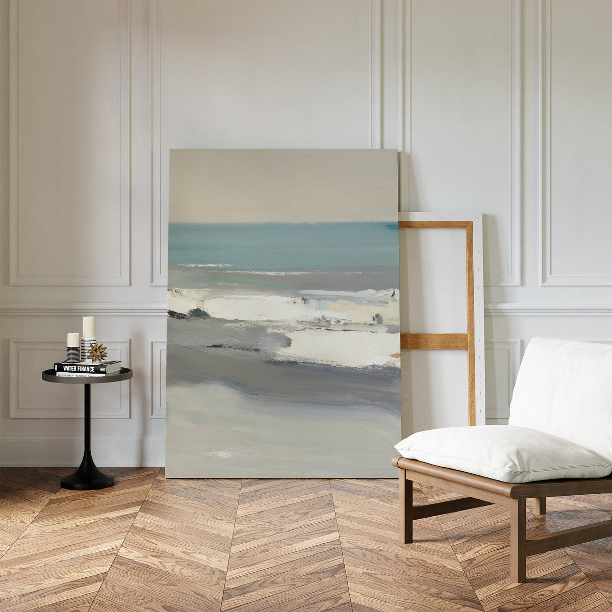 The Sandy Retreat Canvas Art - Vertical leans against a white paneled wall in a minimalist room with herringbone floors, evoking seaside vibes next to a cushioned chair and a round table with candles and books.