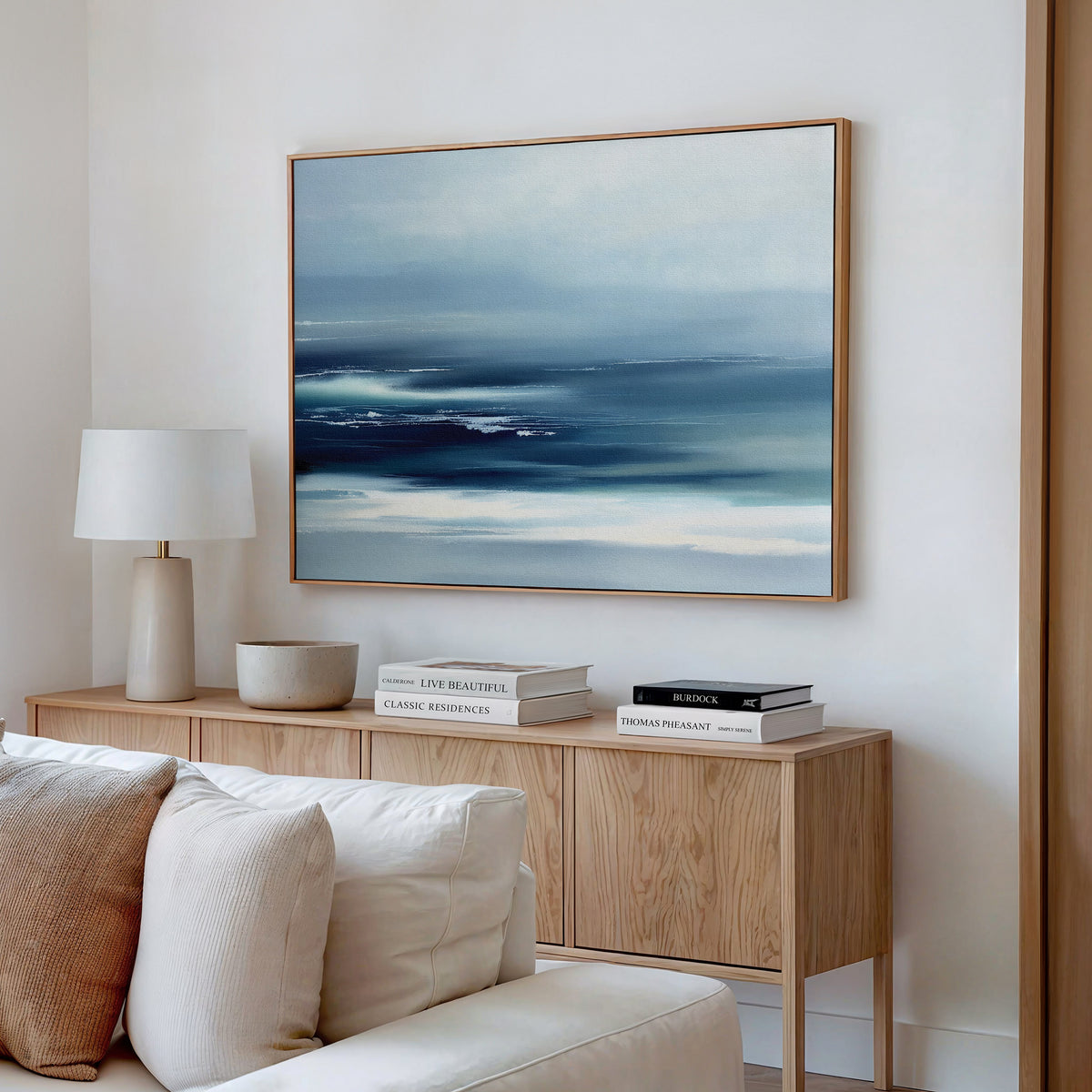 A modern living room corner with a light wooden sideboard, books, a white lamp, a bowl, and the Sapphire Current Canvas Art - Horizontal hanging above a cream sofa.