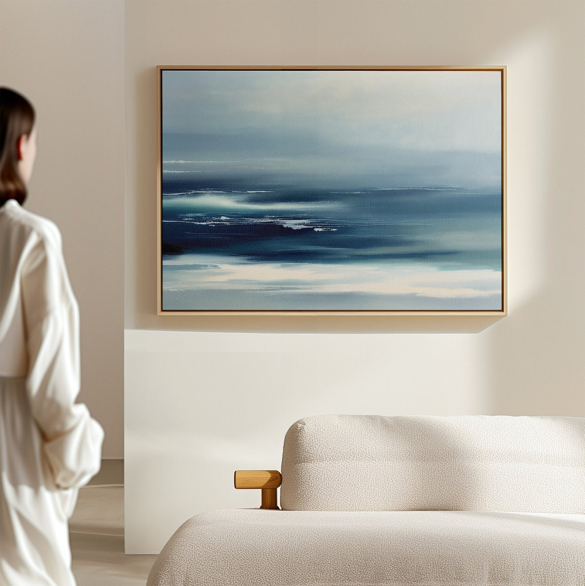 A woman in white stands in a minimalist room, admiring the Sapphire Current Canvas Art - Horizontal, a blue and white abstract print above a light sofa, adding serene Nordic Park vibes.