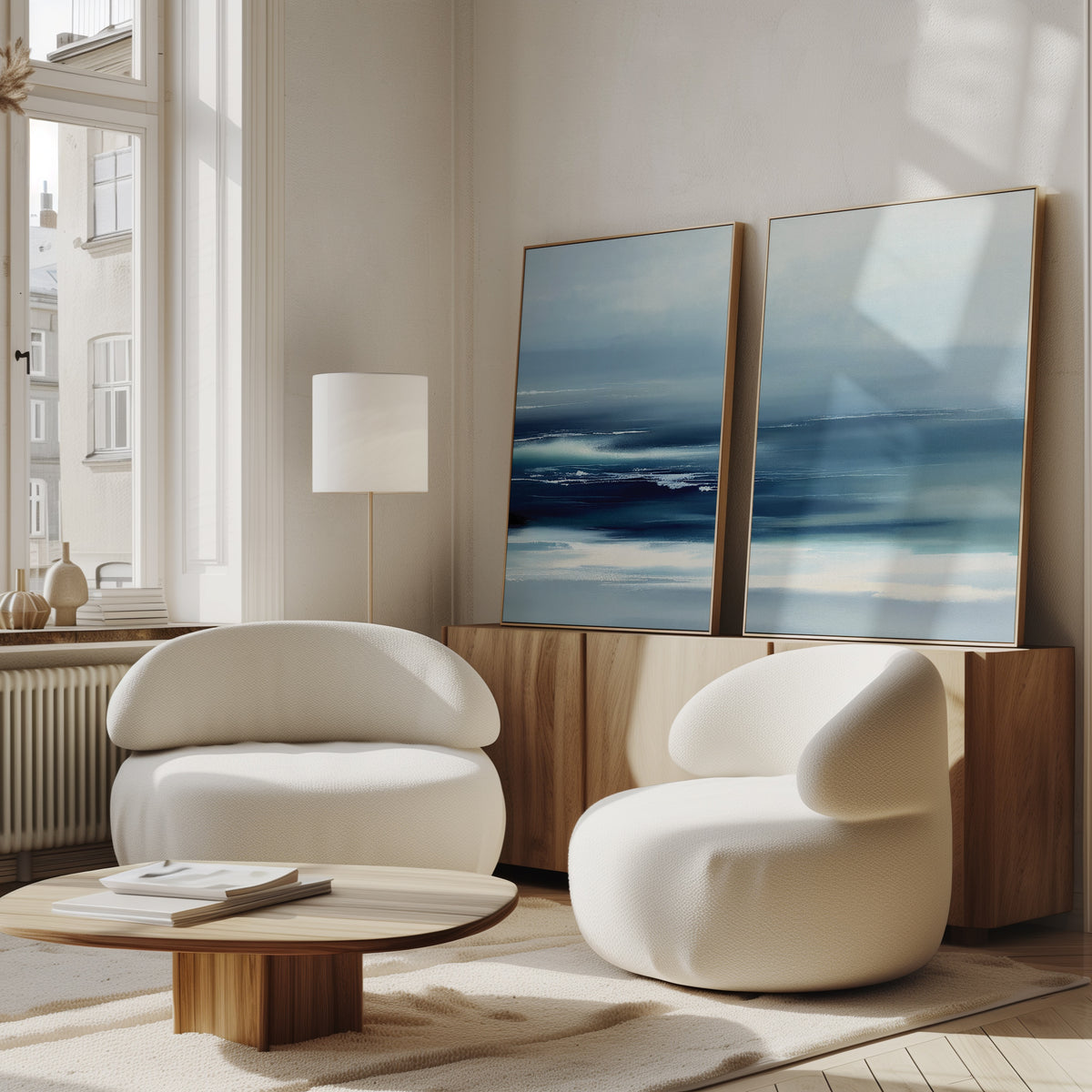 A modern living room featuring two white, curved armchairs, a low wooden coffee table, and a wooden sideboard. The Sapphire Current - Set Of 2 wall art brings color, all aglow from natural light streaming through large windows.
