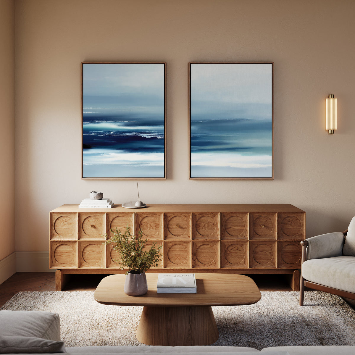 A modern living room features a wooden sideboard, a beige sofa, books on a wooden coffee table, and a small plant. The Sapphire Current - Set Of 2 wall art adds tranquility, while warm lighting creates a cozy atmosphere.