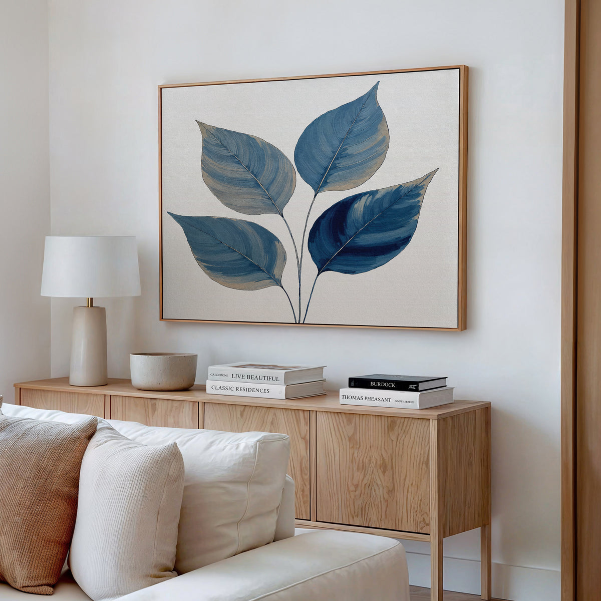 A minimalist living space features a wooden sideboard with books, a lamp, and a ceramic bowl. Above the neutral couch hangs “Sapphire Leaves Canvas Art - Horizontal,” a bold blue botanical print with five large leaves.