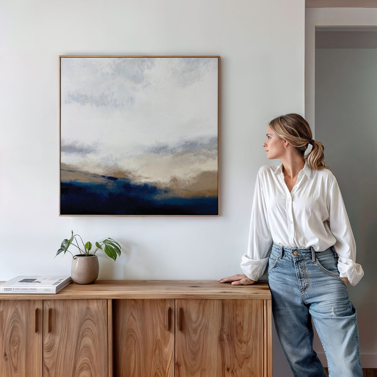 A woman in a white blouse and jeans leans against a wooden cabinet, admiring the Sapphire Tides Canvas Art - Square featuring vibrant cobalt blues above. A small plant and book create a serene, modern living room ambiance.