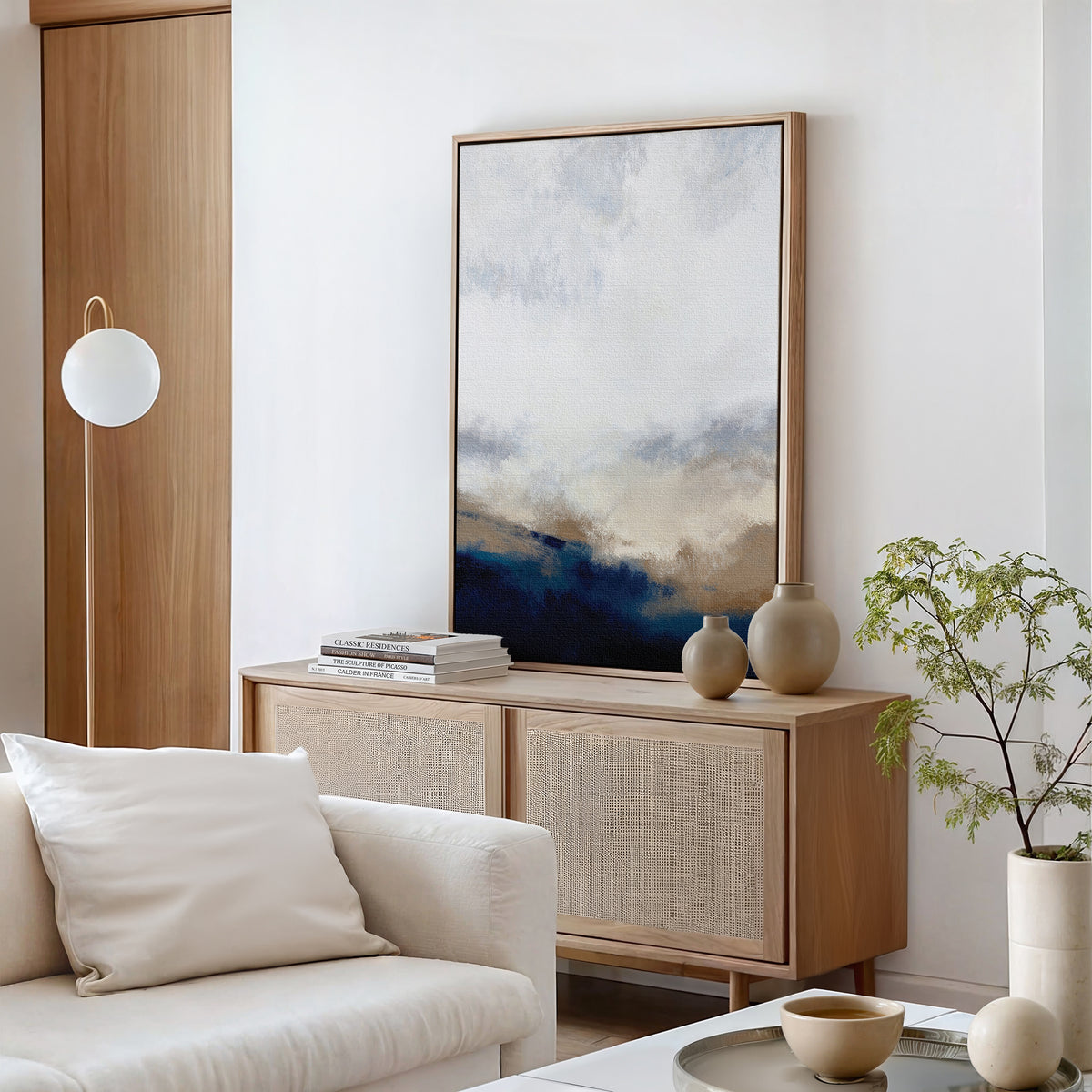 Minimalist living room with a light wood sideboard, Sapphire Tides Canvas Art - Vertical, beige vases, stacked books, a white sofa with cushions, a small plant, and modern hanging globe light. Neutral tones elevate modern style.