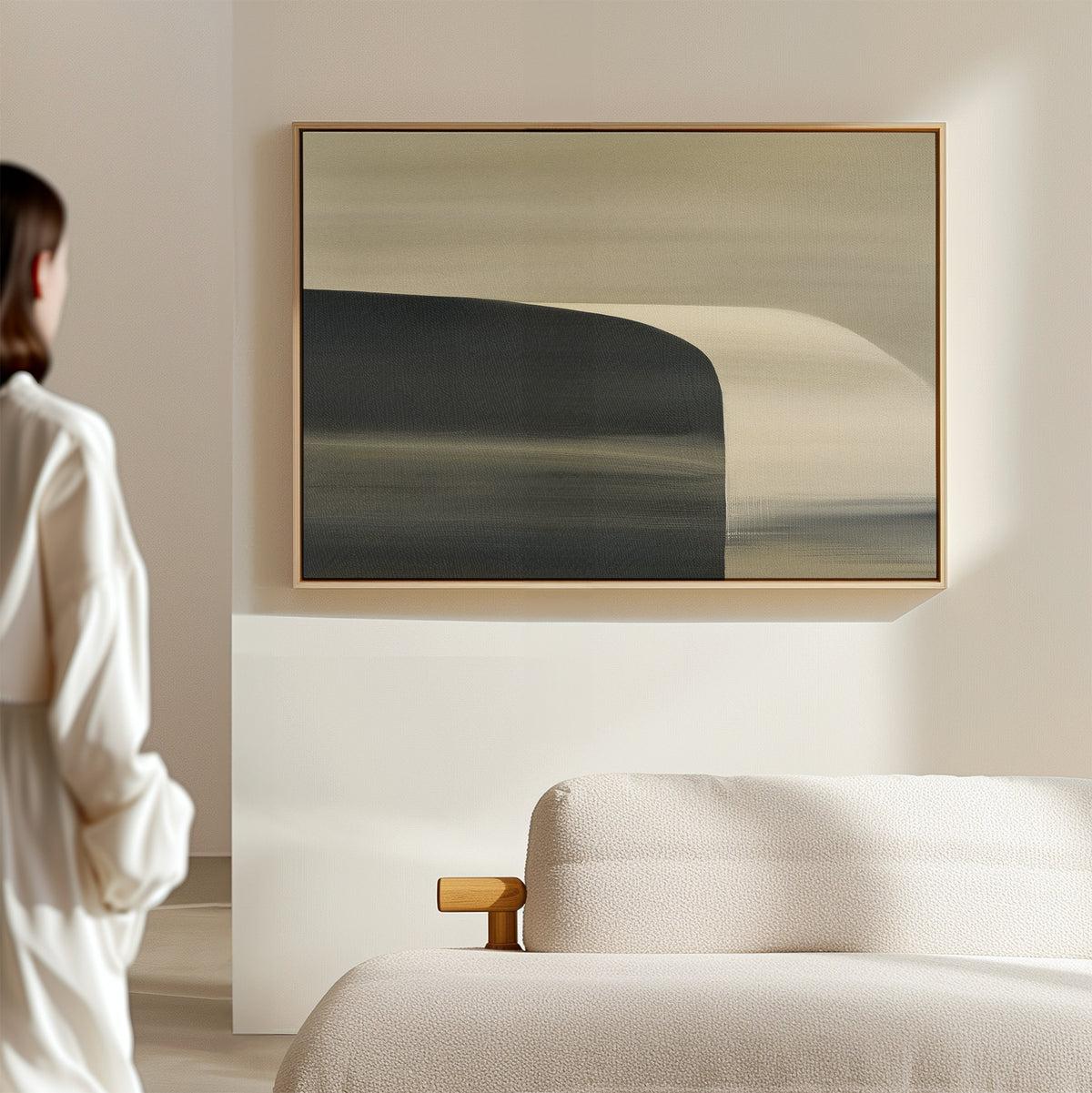 A person in a white outfit admires the Sculptural Forms Canvas Art - Horizontal, featuring soft, muted colors above a beige textured couch in a minimalistic, sunlit living room with modern decor.