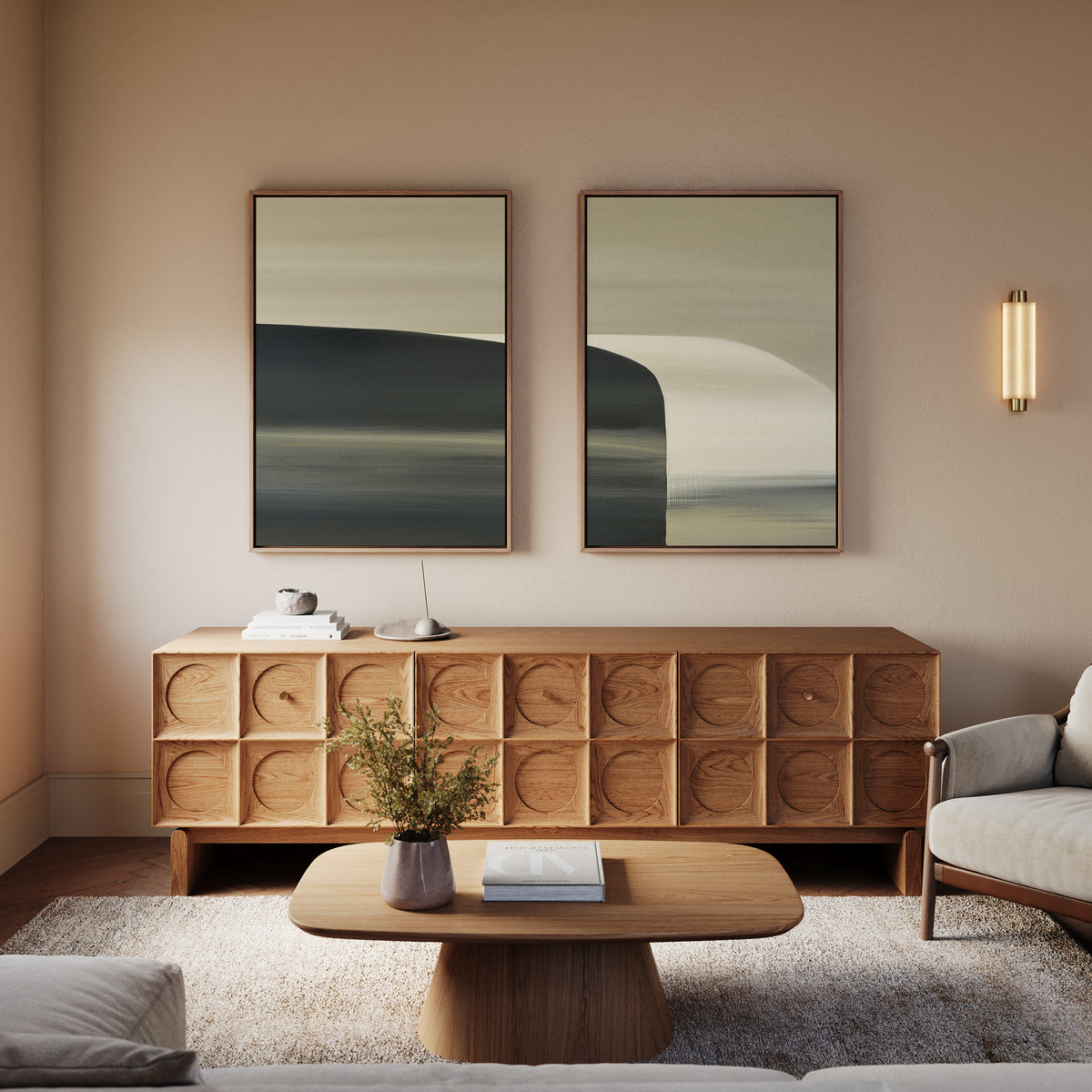 A minimalist living room featuring a wooden coffee table, Sculptural Forms – Set Of 2 atop a textured sideboard, two abstract landscape paintings, a modern wall sconce, and a light-colored sofa with cushion—ideal for fans of modern style.