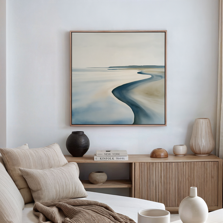 A minimal, coastal-themed living room includes a beige sofa with striped pillows, a wooden sideboard with vases and books, and calming coastal wall art—Serene Coastline Canvas Art—displayed in a frame above the cabinet.