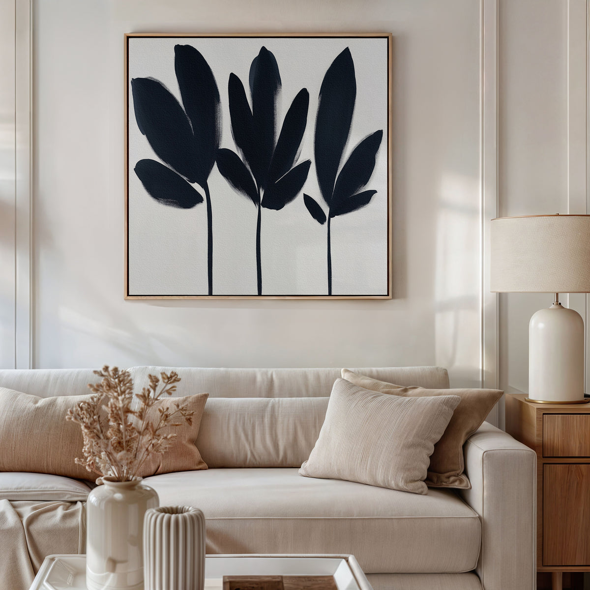 A modern living room with a beige sofa, cream cushions, wooden side table and lamp, vase of dried flowers, and the Shadow Stems Canvas Art - Square with black-and-white botanical silhouettes displayed on the wall.