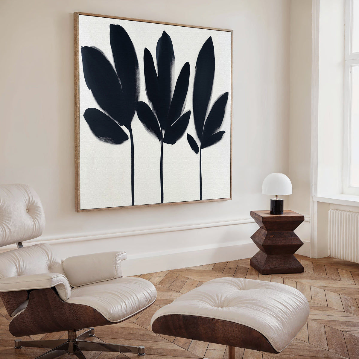 A modern, minimal living room features a white lounge chair with ottoman, wooden side table with a small white lamp, and the Shadow Stems Canvas Art - Square—a large black-and-white botanical print—on the wall.