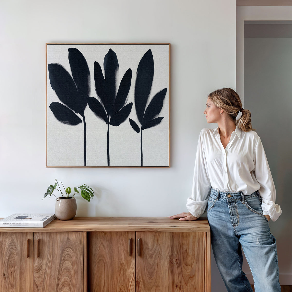 A woman in a white blouse and jeans leans against a wooden cabinet, admiring the Shadow Stems Canvas Art - Square. The minimalist botanical print adds chic style to her modern living room, complemented by a potted plant and a book for serenity.