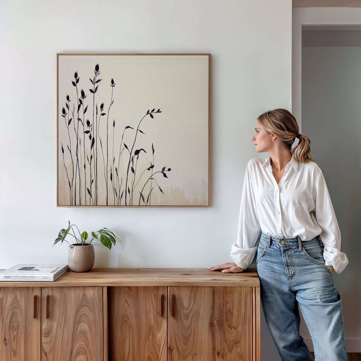 A woman in a white blouse and jeans leans on a wooden cabinet, admiring the Silhouette Meadow Canvas Art - Square. A small potted plant and book nearby enhance the calm Nordic decor of the modern room.