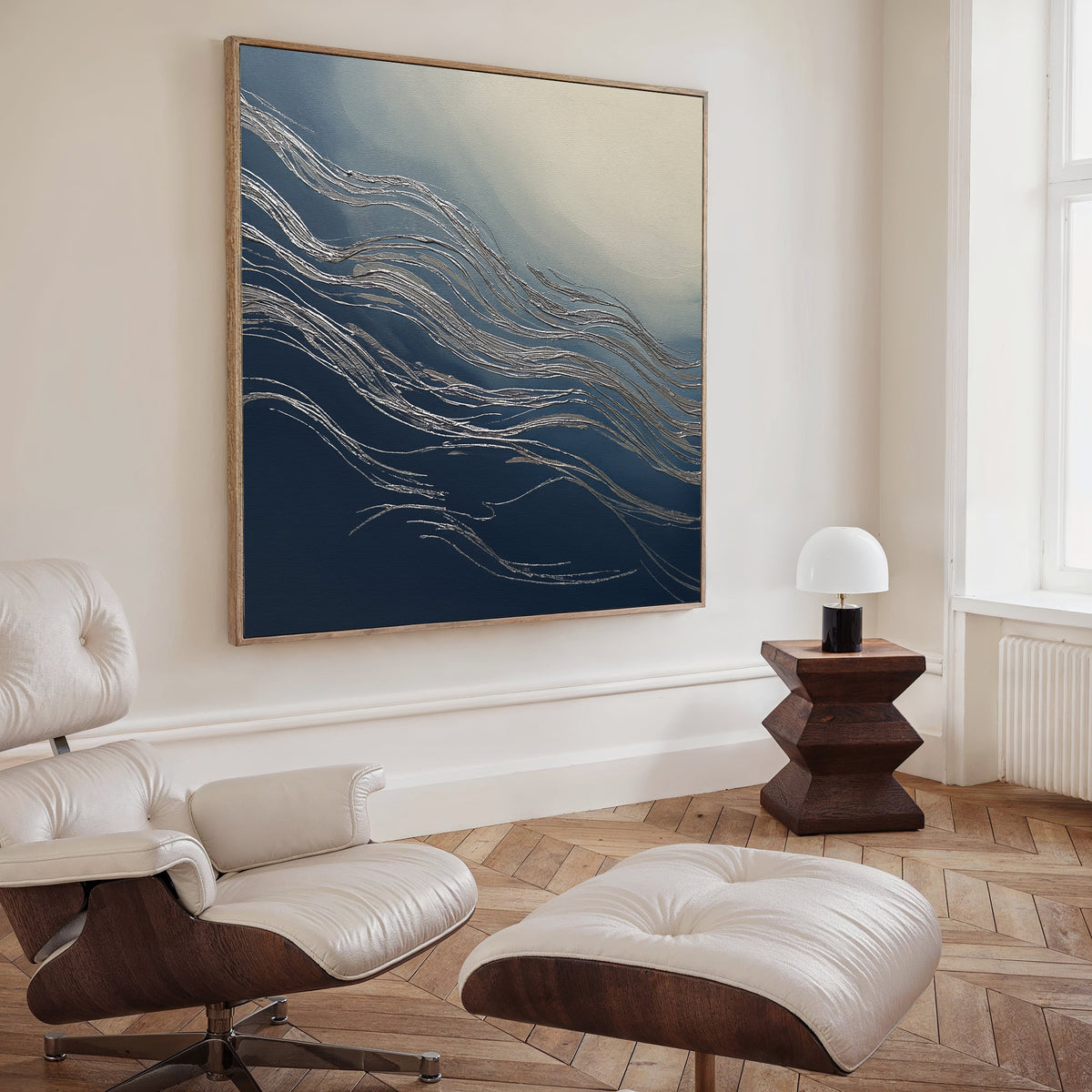 A modern living room features a white leather lounge chair with ottoman, a geometric wooden side table topped by a white lamp, and the Silver Current Canvas Art - Square, whose metallic wavy lines bring contemporary sophistication to the space.