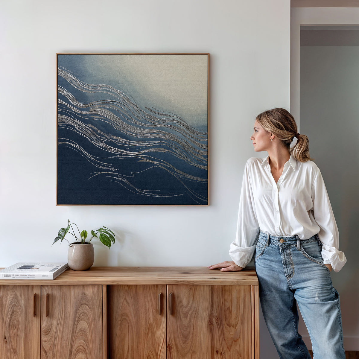 A woman in a white shirt and jeans leans against a wooden sideboard beside the Silver Current Canvas Art - Square, its abstract navy and gold waves adding contemporary sophistication, complemented by wall art, books, and a potted plant.