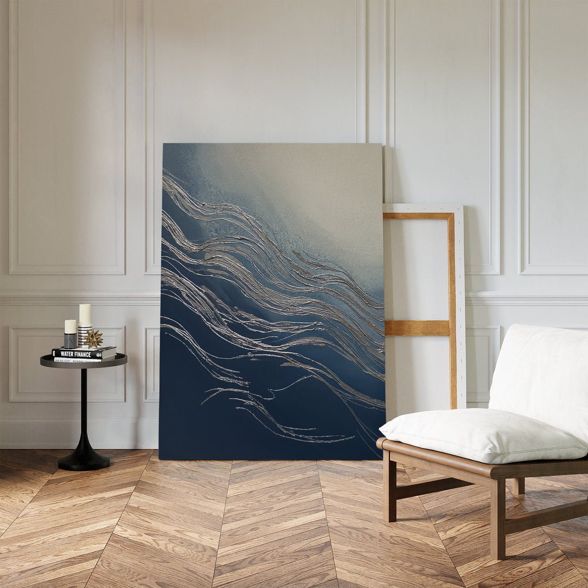 A modern living space with a herringbone wood floor, a white cushioned chair, a sleek black side table with candles and books, and Silver Current Canvas Art - Vertical leaning against a white paneled wall.