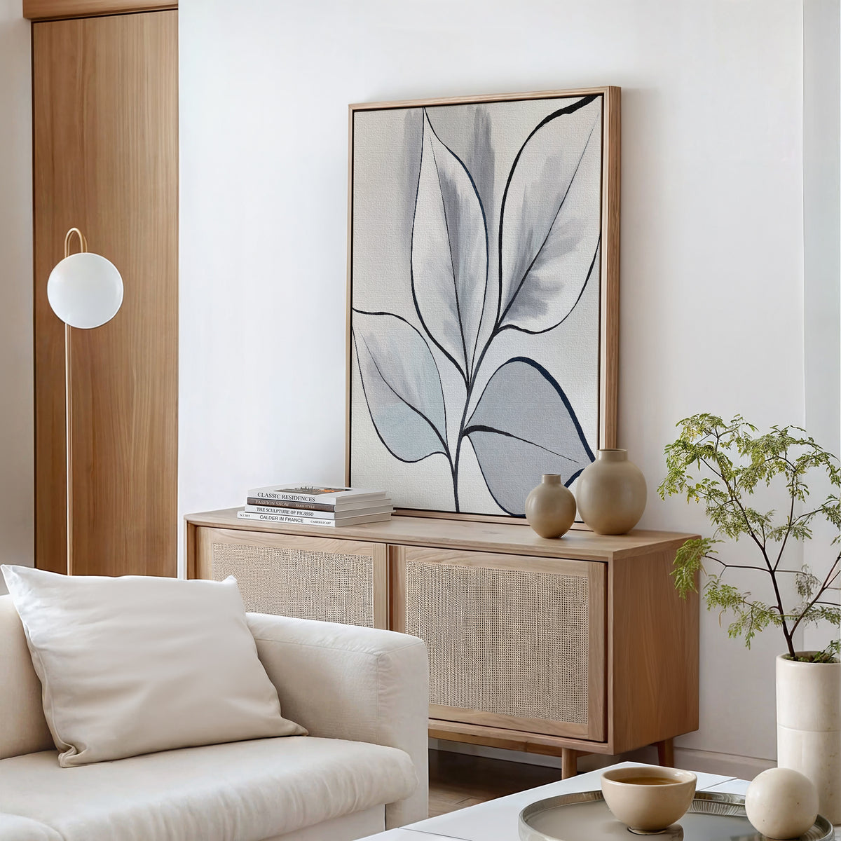 A minimalist living room with a light wooden sideboard, the Silver Leaves Canvas Art - Vertical, neutral vases, a small potted plant, a white sofa, and a modern globe floor lamp creates a bright and calming atmosphere.