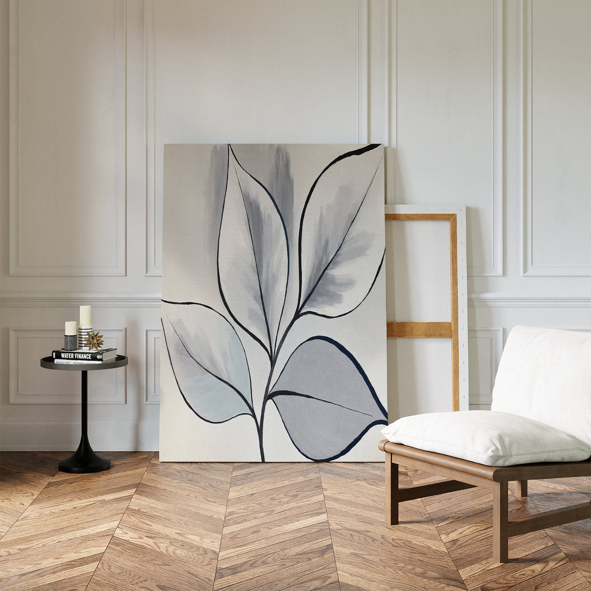 A minimalist room with herringbone wood floors, white paneled walls, a modern white chair, black round side table with candles, and the Silver Leaves Canvas Art - Vertical in muted tones leaning against the wall as decor.