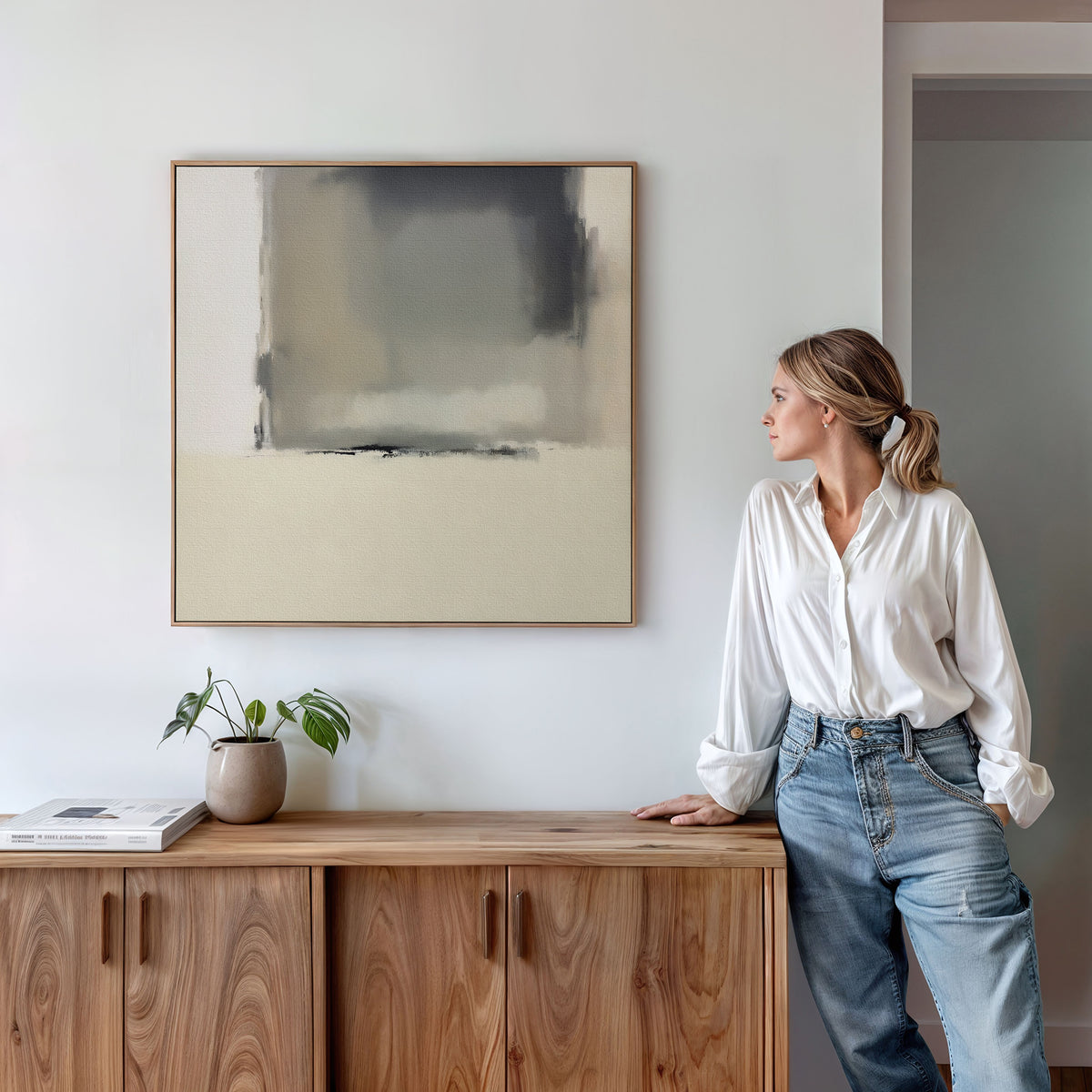 A woman in a white blouse and jeans leans against a wooden sideboard, gazing at the Smoke Boundary Canvas Art - Square. A potted plant and closed book sit nearby in this modern, minimalist room.