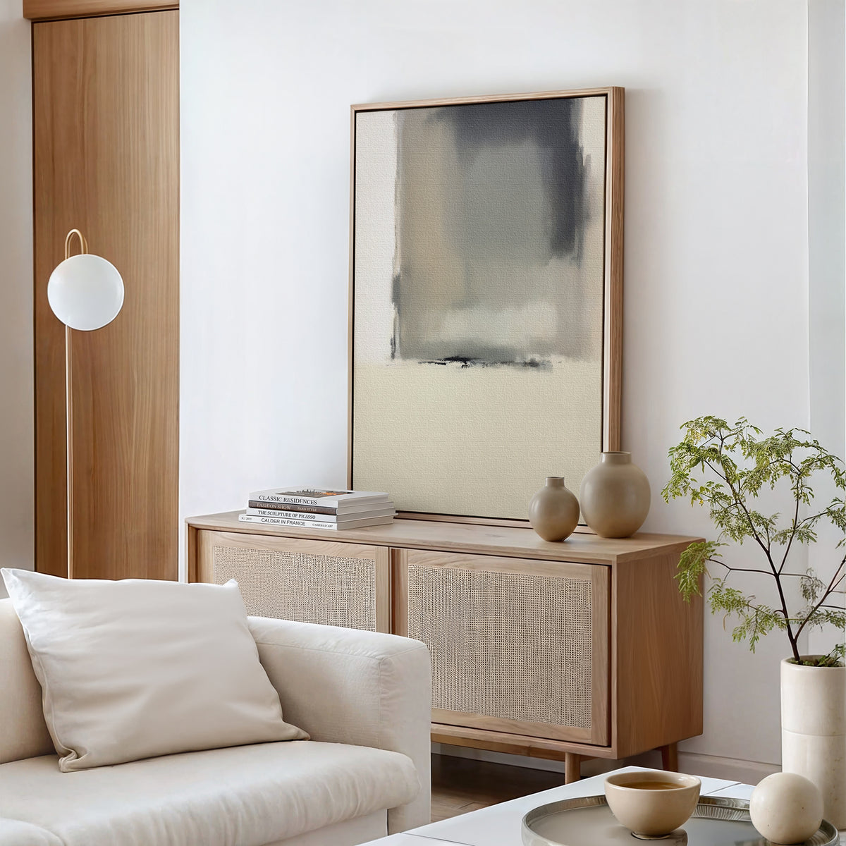 A modern living room with a light wood sideboard, the minimalist Smoke Boundary Canvas Art - Vertical adding neutral elegance, decorative vases, stacked books, a potted plant, a white globe floor lamp, and a white sofa with cushion for serene style.