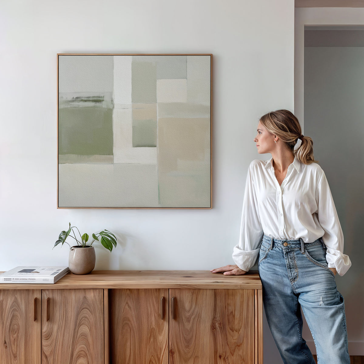 A woman in a white blouse and jeans leans on a wooden cabinet, gazing at the Soft Geometric Planes Canvas Art - Square. A potted plant and books on the cabinet add warmth to the modern, neutral-toned room.
