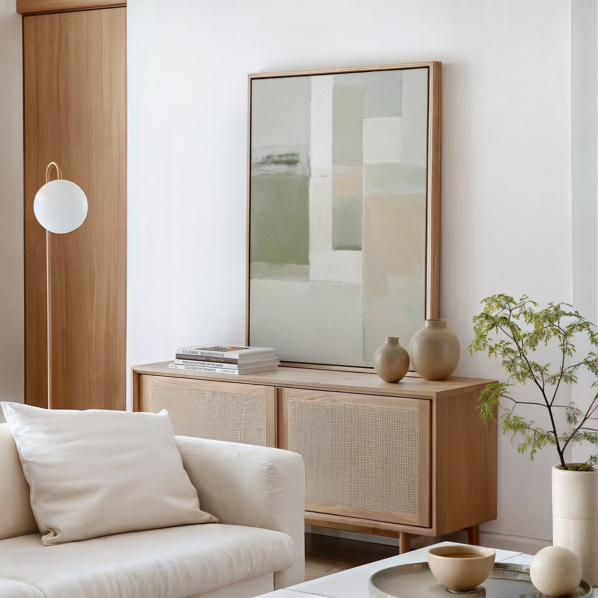 A minimalist living room with a light wood sideboard, white cushioned sofa, neutral vases, stacked books, abstract Soft Geometric Planes Canvas Art - Vertical in sage green and beige tones, plus a round white lamp for a contemporary touch.