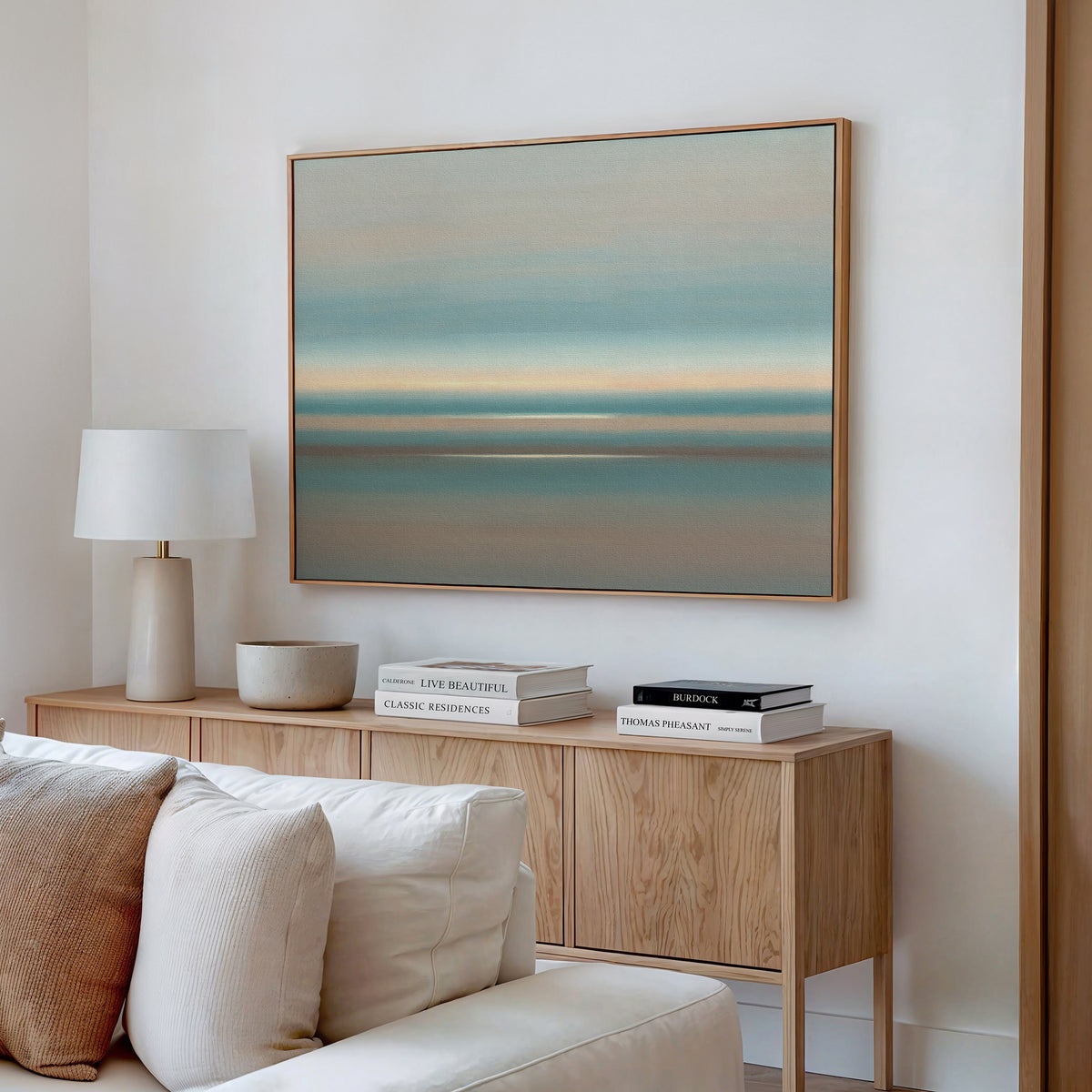 A minimalist living room featuring the Soft Tide Layers Canvas Art - Horizontal in soft blue and beige tones hanging above a cream-colored sofa, paired with a light wood sideboard, stacked books, a lamp, and a bowl.