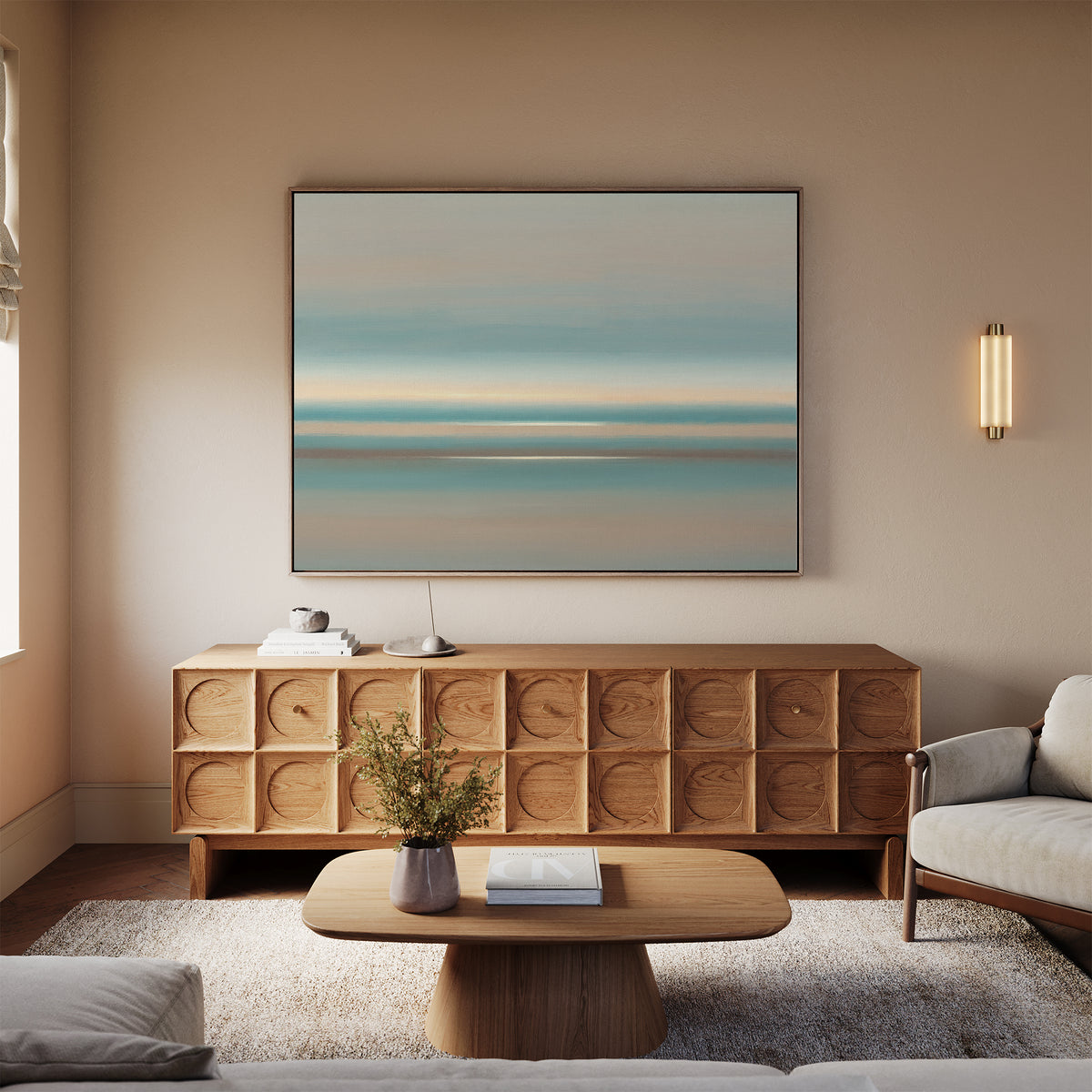 A modern living room features the Soft Tide Layers Canvas Art - Horizontal above a wooden sideboard, with a light wood coffee table, beige sofas, wall sconce, small vase of flowers on a soft rug, and natural light streaming in from the left.