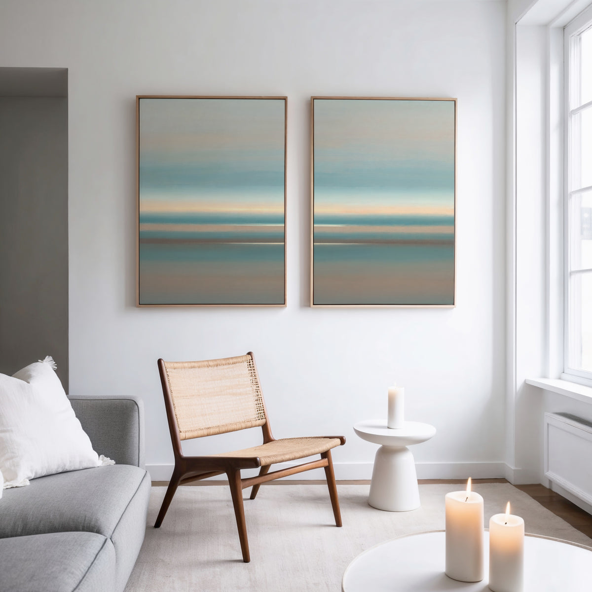A minimalist living room with a gray sofa, wooden chair with woven seat, small white side table, and three lit candles. The "Soft Tide Layers - Set Of 2" coastal wall art hangs above the chair on a white wall, light streaming in.