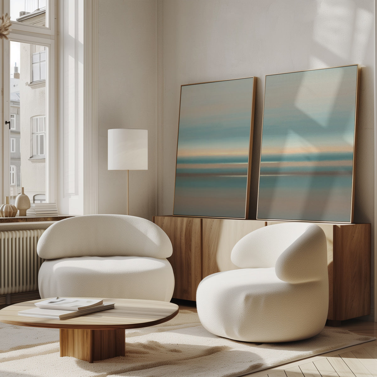 A modern living room with two curved white armchairs, a round wooden coffee table, and Soft Tide Layers - Set Of 2 abstract prints in blue and beige on a wooden cabinet by a tall window. Sunlight fills this serene Nordic Park-inspired space.