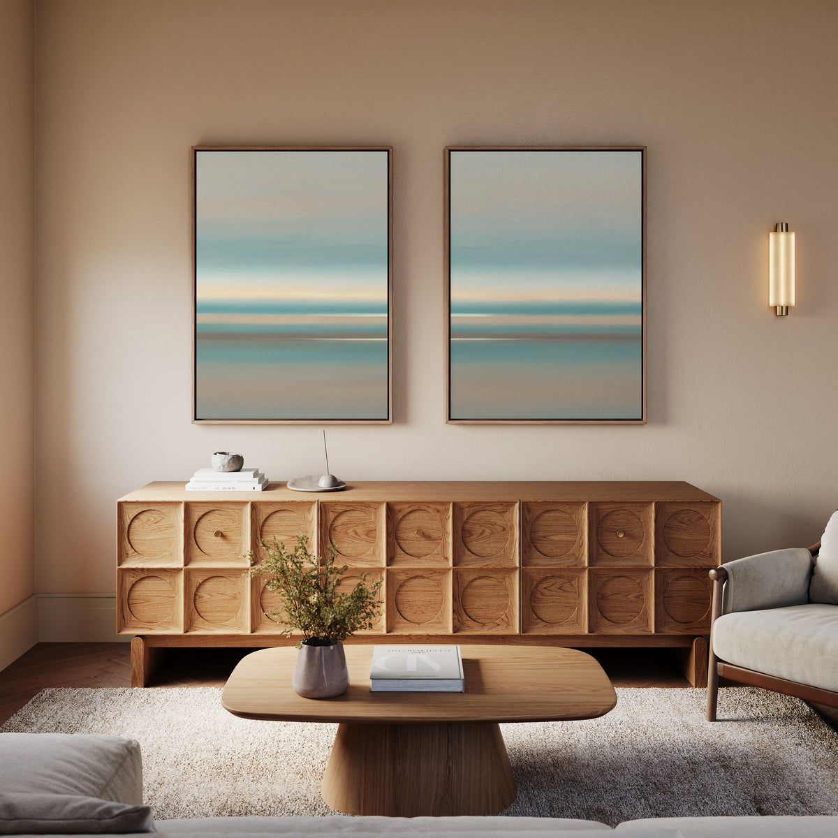 A modern living room with a wooden sideboard, Soft Tide Layers - Set Of 2 wall art in blue and beige above it, a round wooden coffee table with books and a vase, and a light armchair on a rug. Warm lighting adds coziness.