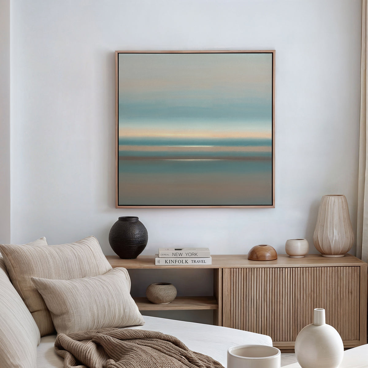 A minimalist living room with a beige sofa, neutral cushions, a wooden sideboard, and the Soft Tide Layers Canvas Art - Square in blue and tan tones above. Decorative vases and books adorn the sideboard.