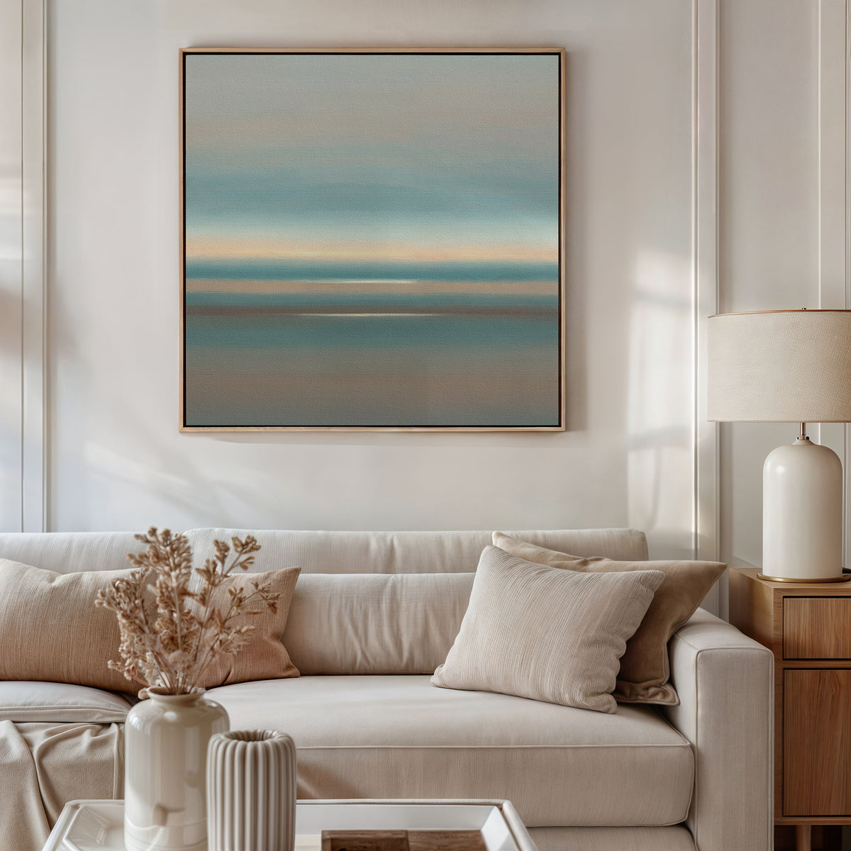 A modern living room with a beige sofa, neutral cushions, a lamp on a wooden side table, dried flowers in a vase, and the Soft Tide Layers Canvas Art - Square featuring calming blue and peach tones adorning the wall.