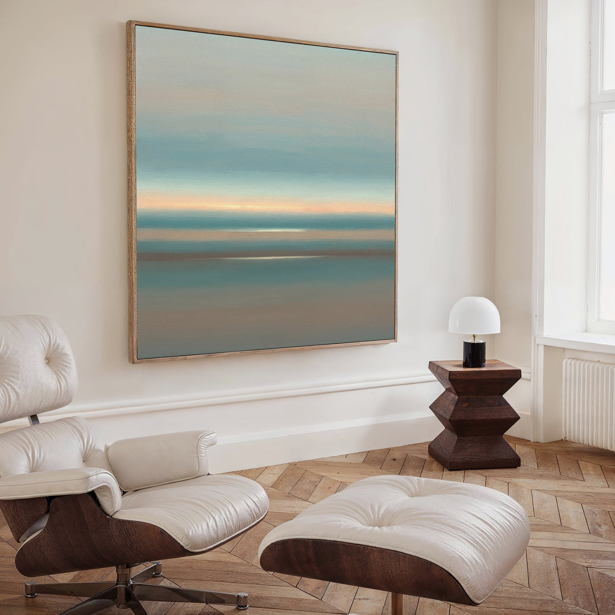 A modern, minimalist living room featuring a white leather lounge chair and ottoman, a geometric wooden side table with lamp, and Soft Tide Layers Canvas Art - Square in soothing blue, green, and orange tones adorning the wall.
